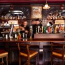 Carved wooden Aulde Dubliner bar with two bartenders mixing drinks beneath rows of liquor bottles and beer taps.