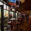 Pub dining room with wooden tables and stools near an open patio door, a mounted TV in the foreground, and guests seated throughout.