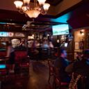 Pub interior with a crystal chandelier, stained glass accents, and a wall-mounted TV, with guests seated at the bar and tables.
