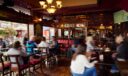 Pub interior with a crystal chandelier and stained glass, guests seated at tables and booths near open doors leading out to a flower-lined patio street.