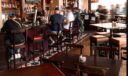 Guests seated at the bar and wooden tables in a pub room with checkered tile flooring and shelves of liquor bottles.