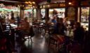 Sunlit pub dining room with 'Stouts' and 'Spirits' stained glass signage, guests at tables, and a view of a flower-lined street outside.