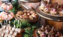 Assorted catering spread featuring shrimp cocktail, melted cheese sliders, and roast beef tartlets on tiered stands.