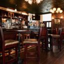 Traditional Irish pub interior with a polished wood bar, high-top tables, ornate tin ceiling, stone walls, and warm globe lighting.