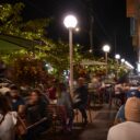 Outdoor patio at night with diners seated beneath string lights and glowing globe lamps along a lively streetscape.