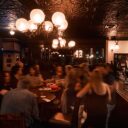 Guests gathered at round wooden tables under an ornate globe chandelier in a busy pub with pressed-tin ceiling and dark wood paneling.