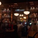 Bar area with 'Heart and Crown' sign and lit Guinness Extra Stout window display, flanked by shelves of liquor bottles and patrons in the foreground.