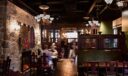 Wide view of a crowded, warmly lit pub interior with pressed-tin ceiling, globe chandeliers, and a long wooden bar.