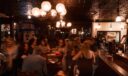 Wide view of a crowded, warmly lit pub interior with pressed-tin ceiling, globe chandeliers, and a long wooden bar.