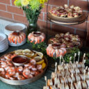 Catering spread with shrimp cocktail, sliced pinwheel wraps, melted cheese toasts, and fresh flowers on a table against a brick wall.