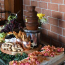 Dessert table with a chocolate fondue fountain, ladyfinger cookies, fresh fruit, cheese, and crackers, decorated with fresh flowers.
